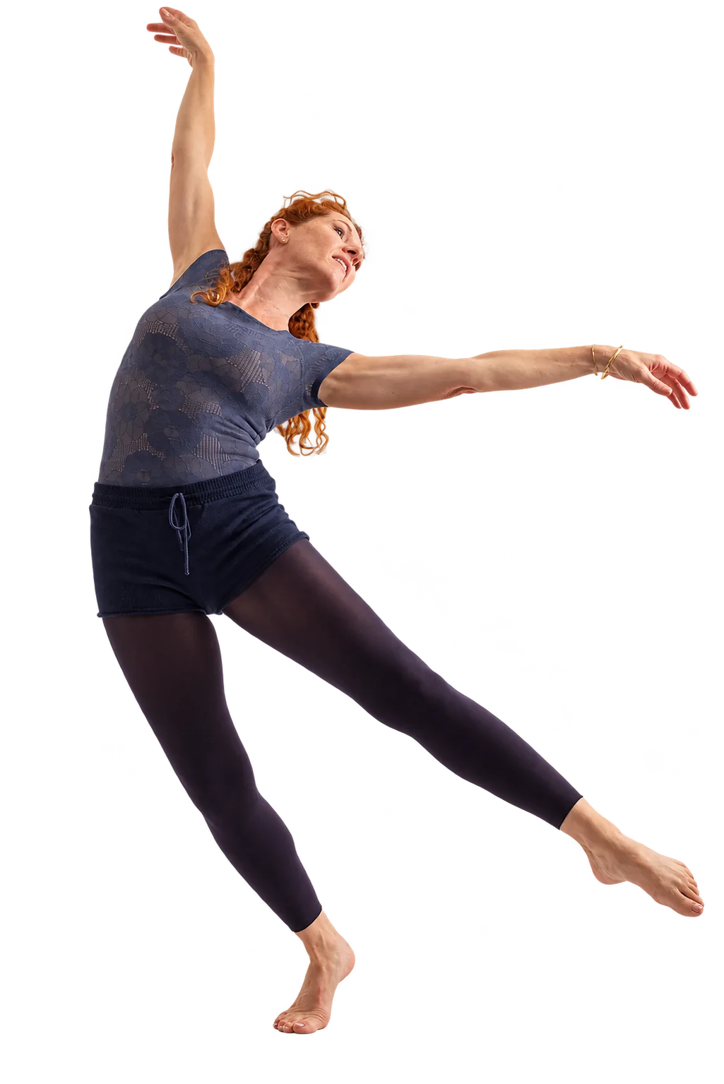 Une danseuse aux cheveux roux, en justaucorps gris-bleu, short sombre et collants, effectue un mouvement gracieux, un bras levé et une jambe tendue, sur fond blanc.
