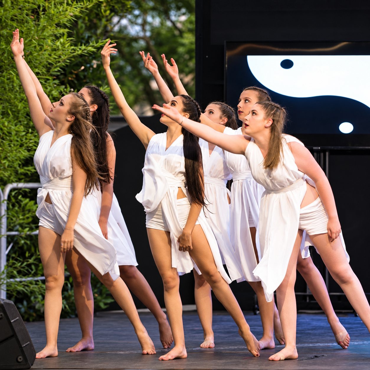 Un groupe de danseuses pieds nus, vêtues de robes blanches, lèvent le bras en chœur sur une scène en plein air devant un écran.