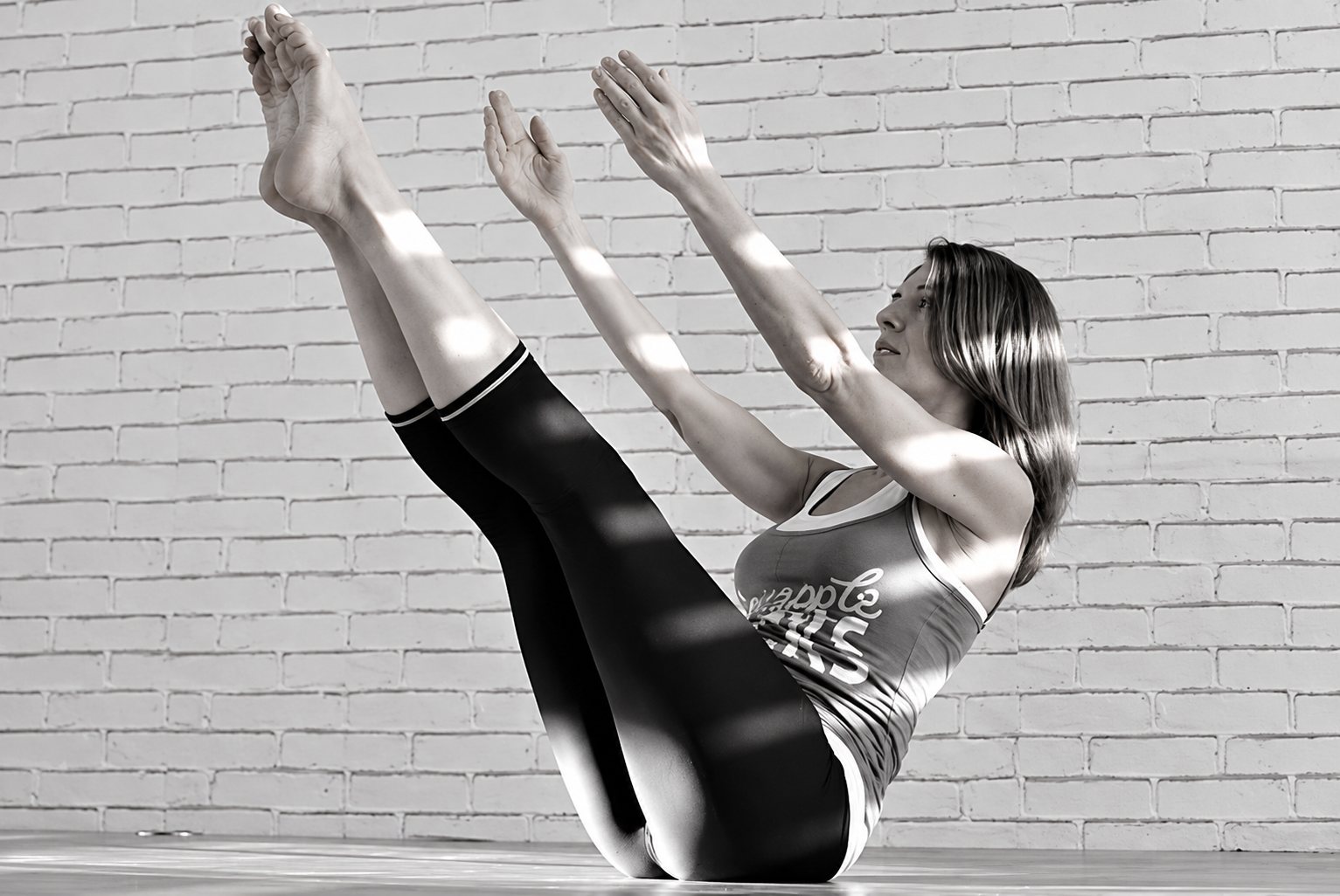 Photo en noir et blanc d'une femme en tenue de sport qui effectue un exercice d'équilibre de Pilates, jambes et bras tendus, devant un mur de briques blanches.