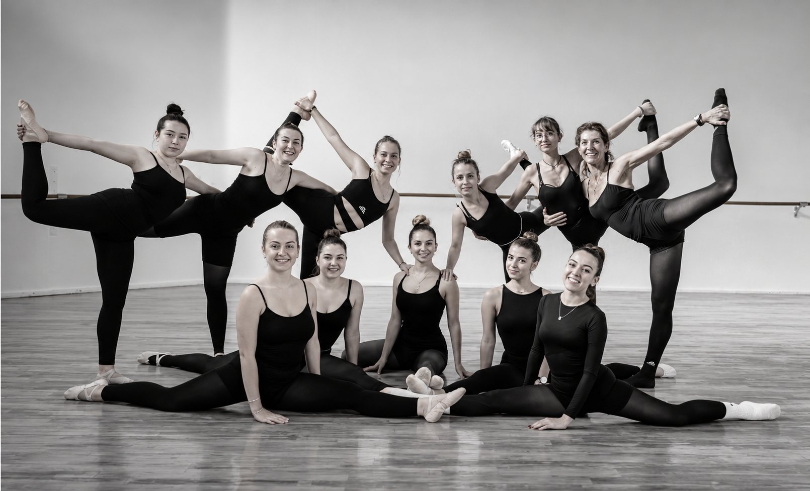 Photo en noir et blanc d'un groupe de danseuses en tenue noire posant ensemble dans un studio de danse, certaines jambes levées, d'autres assises au sol.