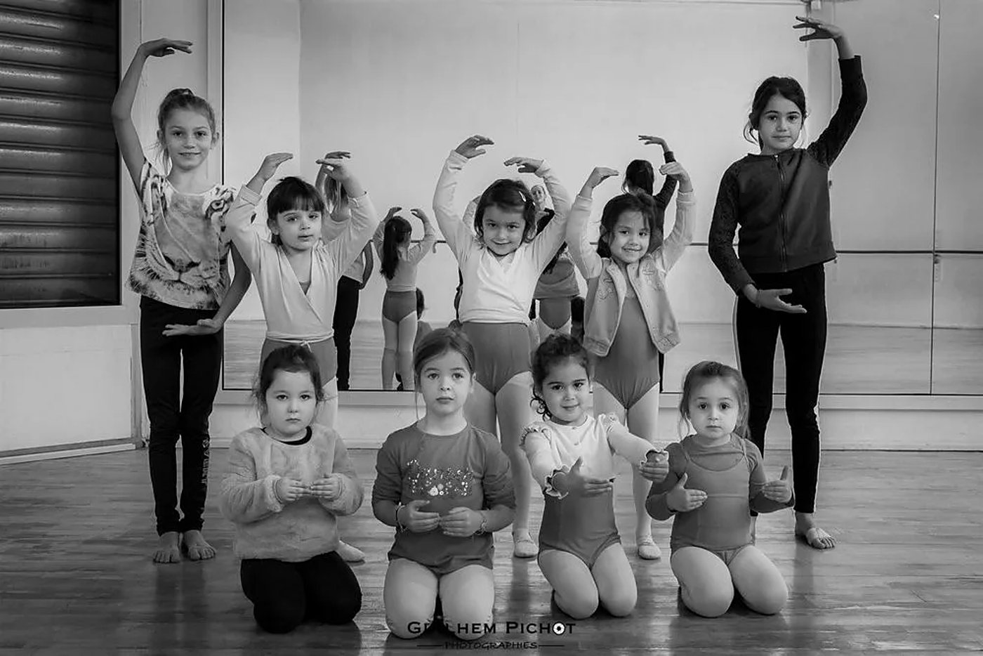 Photo en noir et blanc de huit petites filles en tenue de danse, les bras en position de ballet, devant un grand miroir dans une salle de danse.