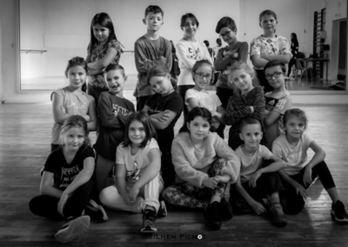 Photo en noir et blanc d'un groupe d'enfants souriants, certains assis au sol et d'autres debout, posant dans une grande salle avec un miroir et un parquet.