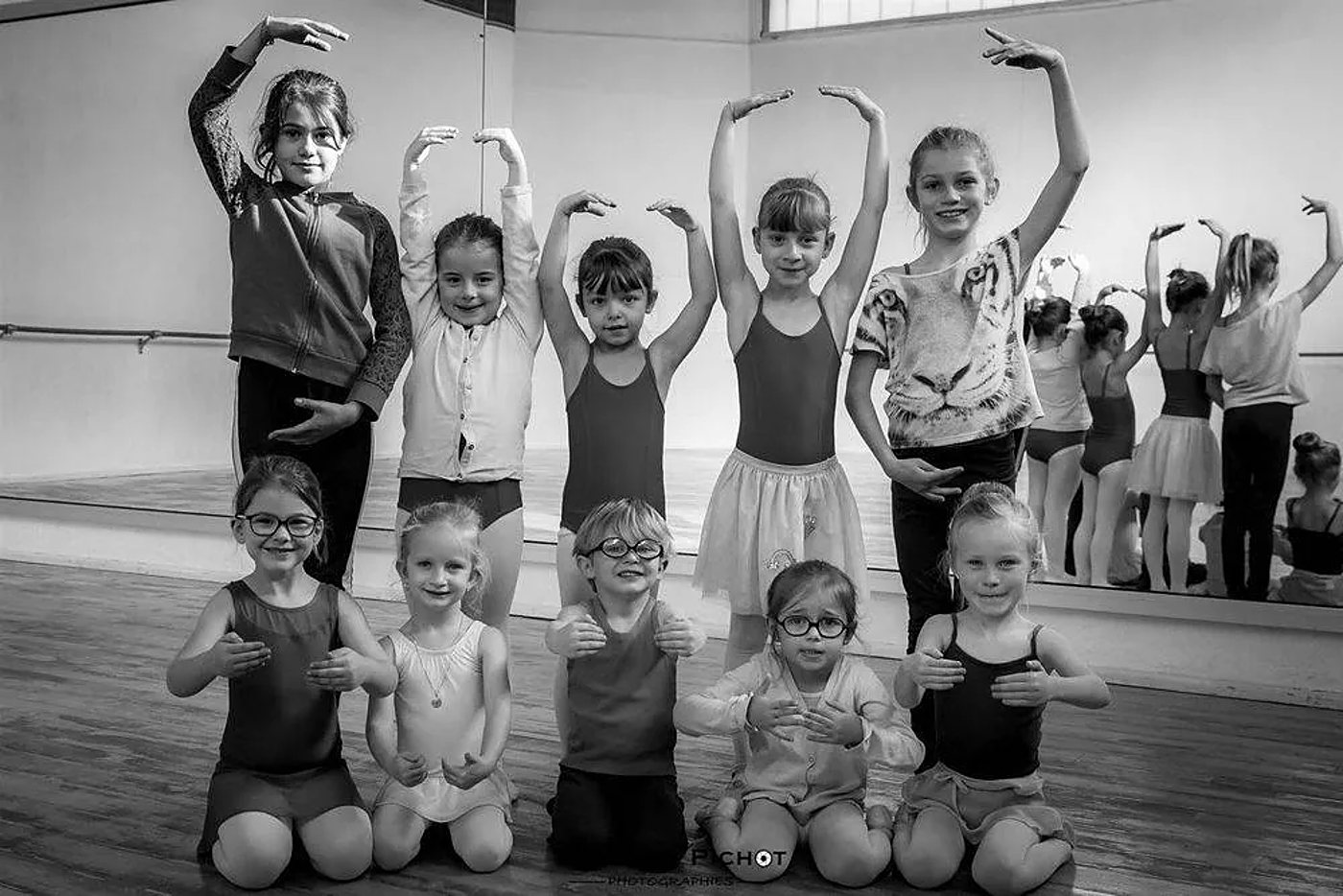 Photo en noir et blanc de dix jeunes danseuses souriantes, bras levés ou accroupies, posant dans un studio de danse devant un miroir.