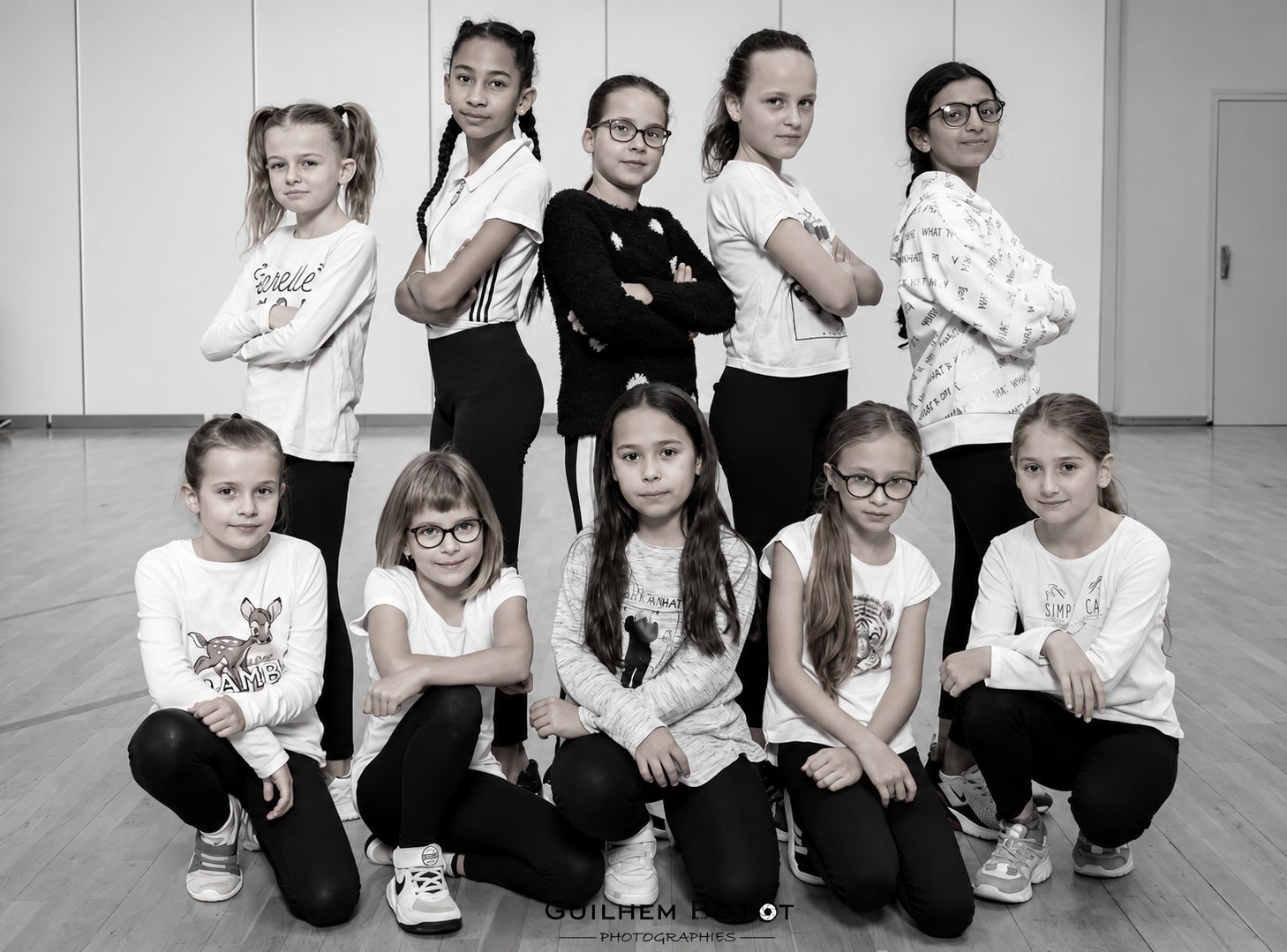 Photo en noir et blanc de dix jeunes filles posant en deux rangées, bras croisés, dans une salle de danse au parquet.