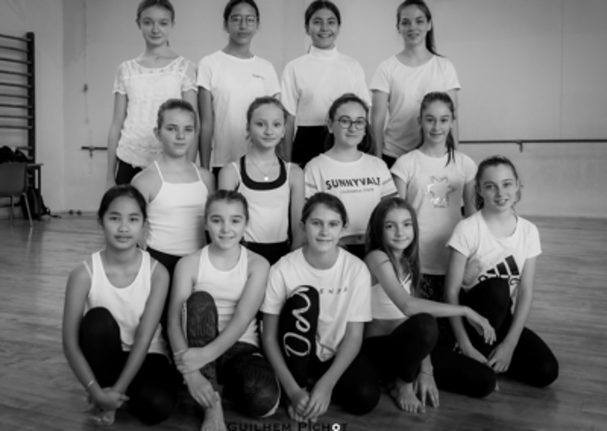 Photo en noir et blanc d'un groupe de jeunes filles en tenue de danse posant sur trois rangs dans une salle au parquet, près d'espaliers et d'une barre.