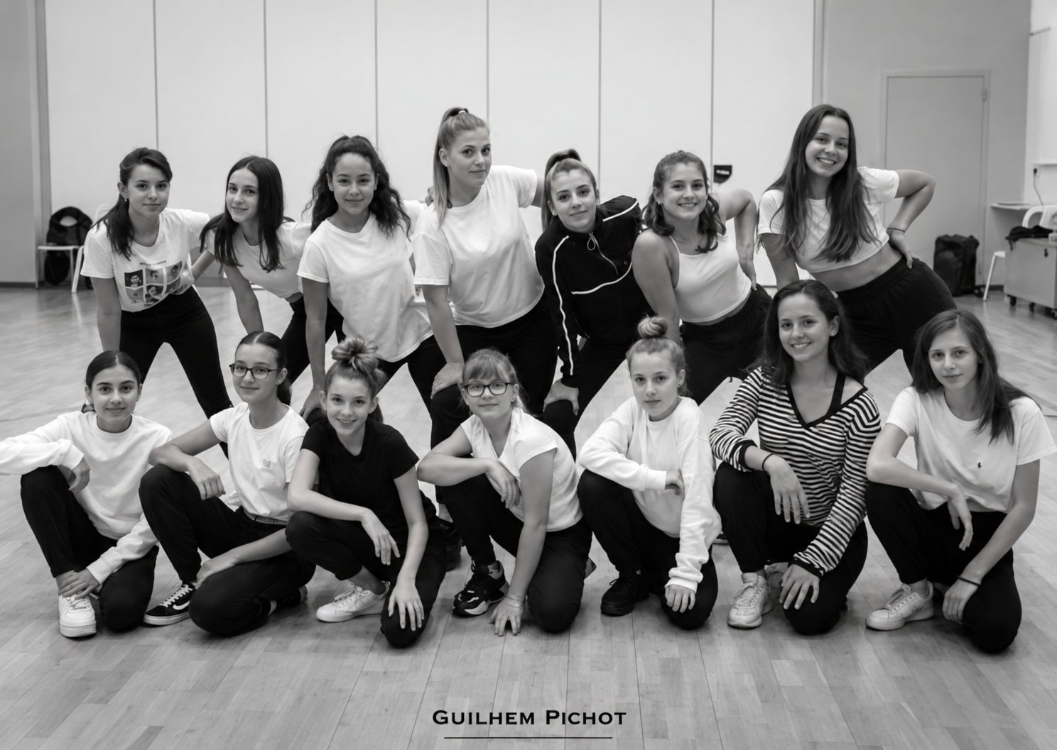 Photo en noir et blanc d'un groupe de jeunes danseuses souriantes, debout et accroupies, posant ensemble dans un studio de danse au parquet clair.