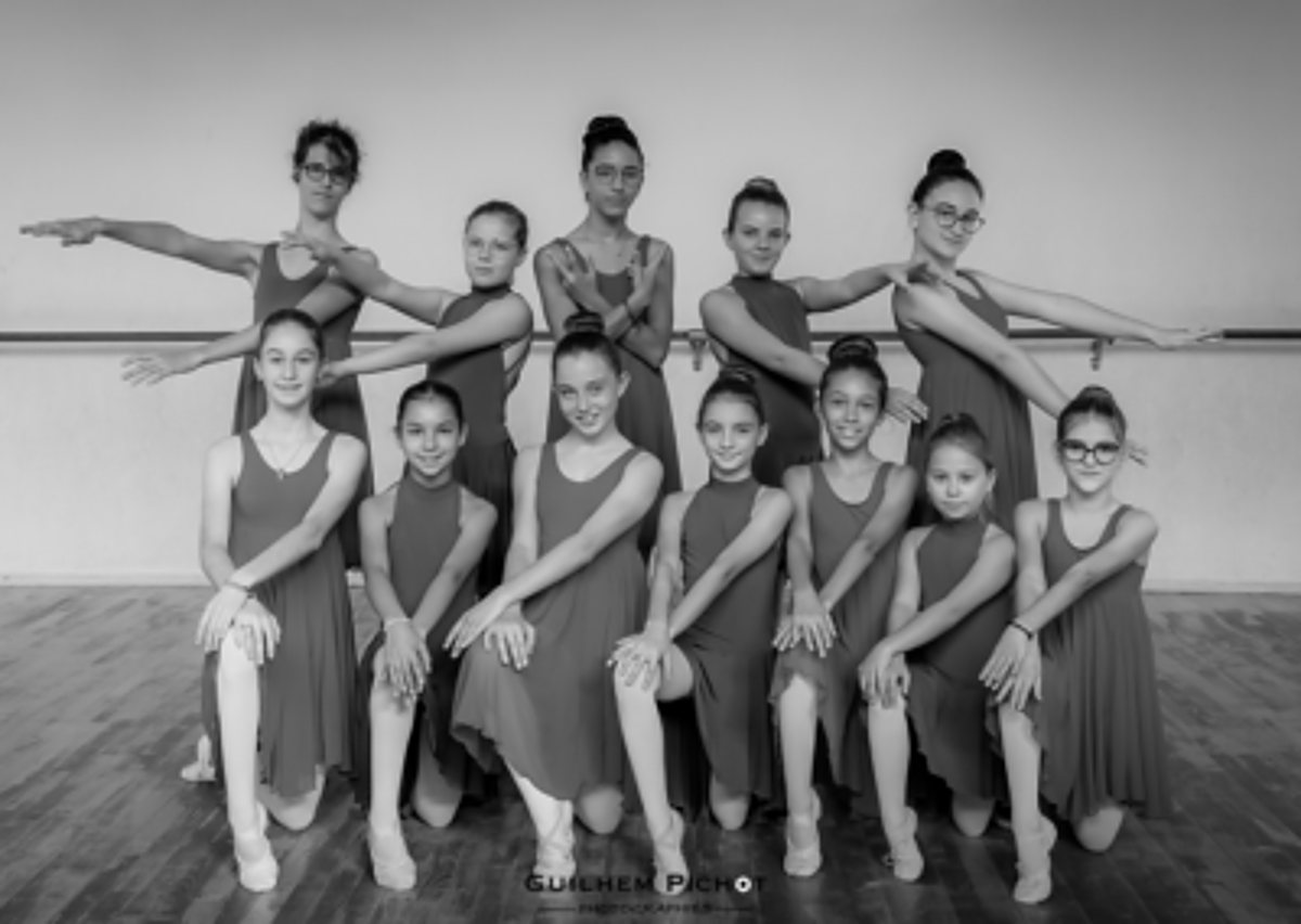 Photo en noir et blanc d'un groupe de jeunes danseuses en robes identiques, posées sur deux rangs dans un studio de danse devant une barre.