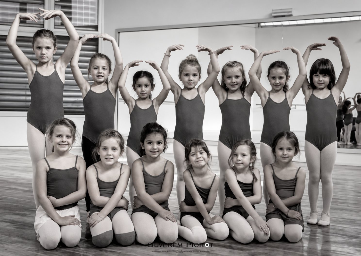 Photo en noir et blanc d'un groupe de jeunes filles en justaucorps posant en deux rangées dans un studio de danse devant un miroir.
