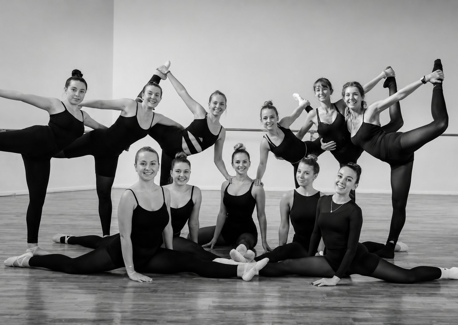 Photo en noir et blanc d'un groupe de jeunes danseuses en tenue sombre qui posent ensemble dans un studio, certaines assises en grand écart et d'autres debout, la jambe levée.