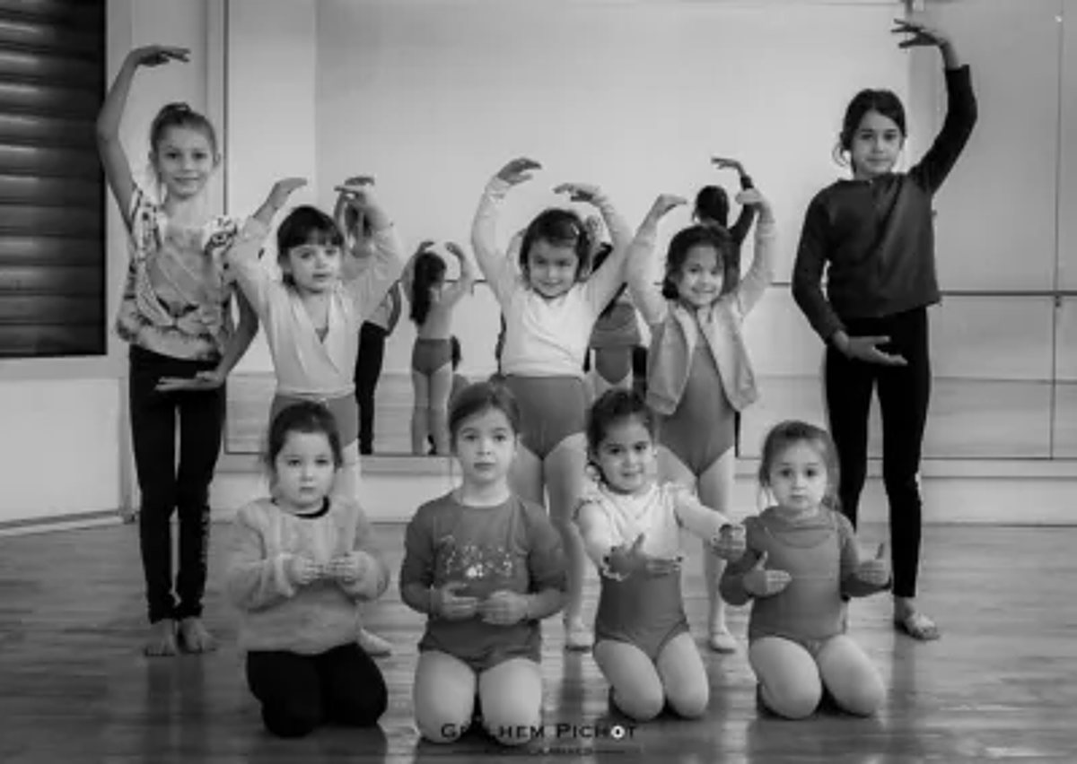 Photo en noir et blanc d'un groupe de petites filles posant dans un studio de danse, certaines à genoux au premier plan et d'autres debout les bras arrondis au-dessus de la tête.