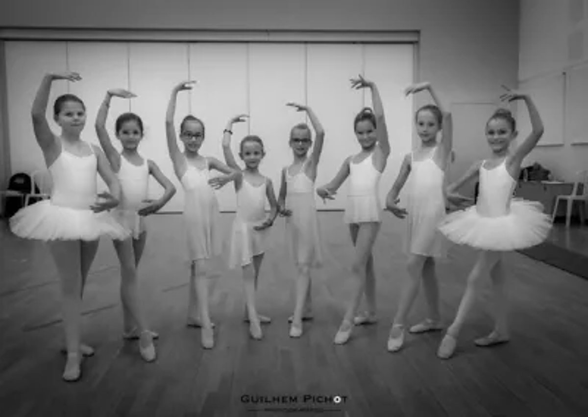 Photo en noir et blanc d'un groupe de neuf jeunes filles en tenue de danse classique, un bras levé, posant dans une salle de danse.