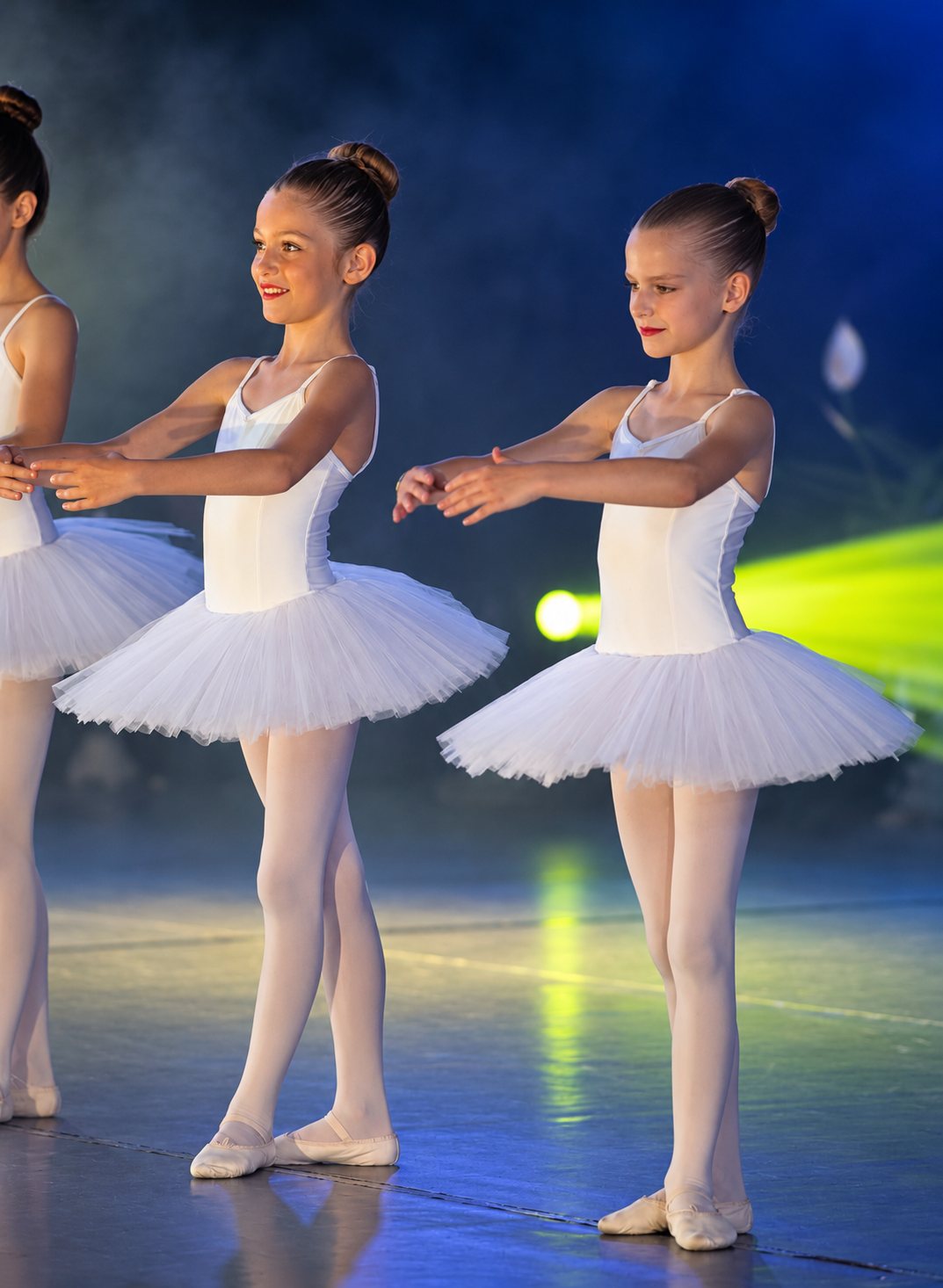 Deux jeunes danseuses en justaucorps et tutu blancs posent les bras tendus sur une scène éclairée de lumières colorées, une troisième élève étant partiellement visible à gauche.