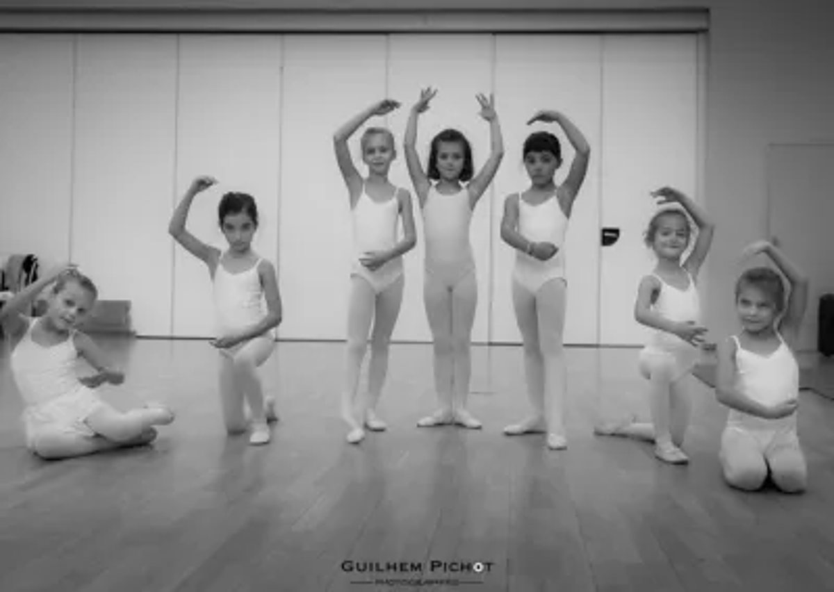 Sept jeunes enfants en tenue de danse classique prennent la pose dans un studio au parquet, photographiés en noir et blanc.