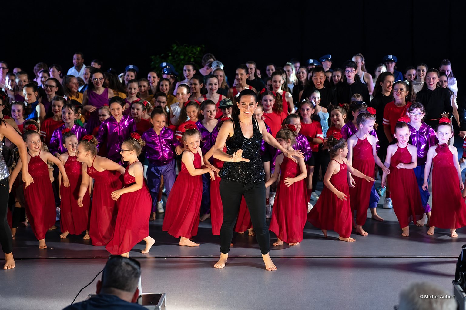 Une professeure de danse en haut noir à paillettes pose pieds nus sur scène, entourée de nombreux enfants en robes rouges et vestes violettes lors d'un spectacle.