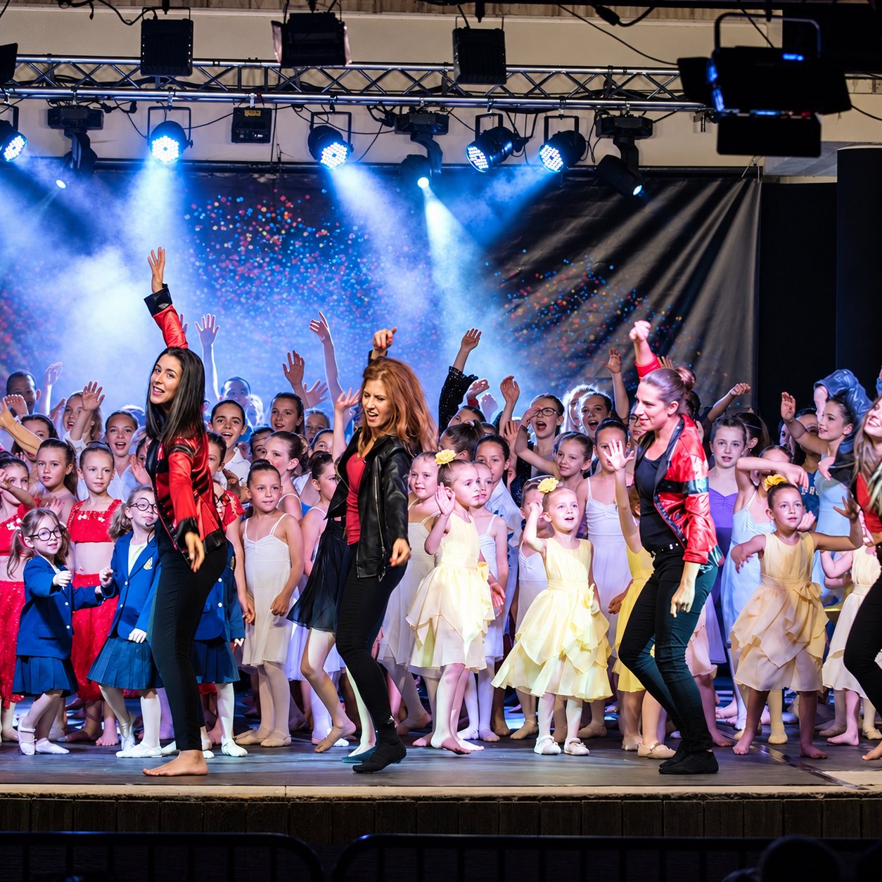 Trois professeures de danse en blouson rouge ou noir dansent devant un groupe d'enfants en tenues de scène, sous des projecteurs bleus et des confettis.